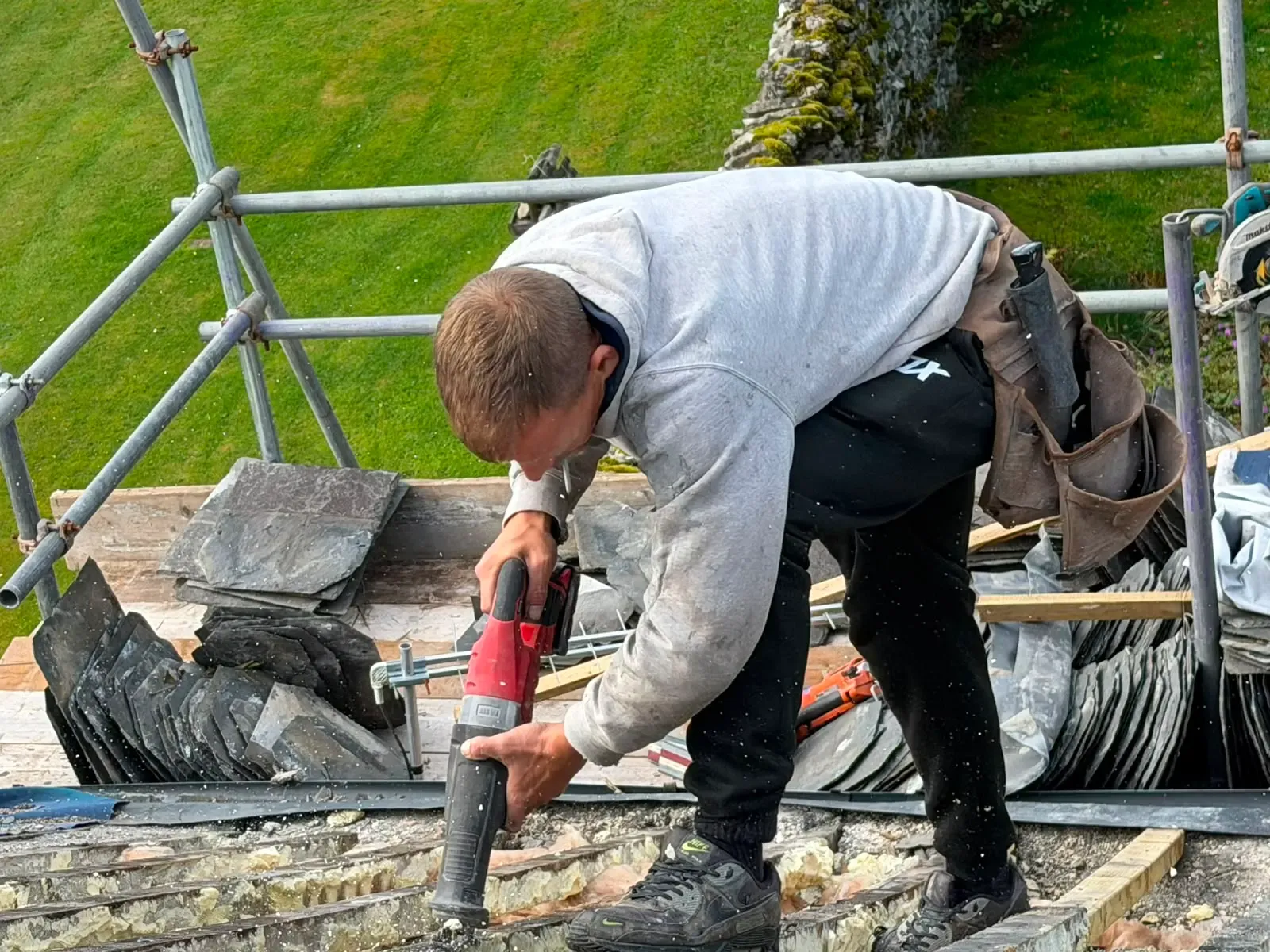 Roofer removing spray foam insulation from rafters and roof battens on a scaffolding platform with slates