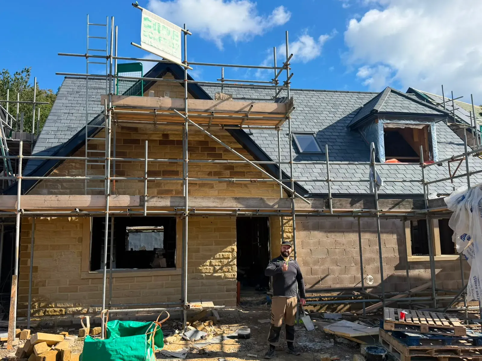 Roofer standing in front of a newly completed slate pitched roof installation on a stone property with scaffolding